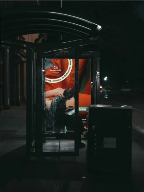 Un homme est assis dans un abribus la nuit, regardant son téléphone. Une publicité lumineuse pour 'Le transit' est visible à l'intérieur.