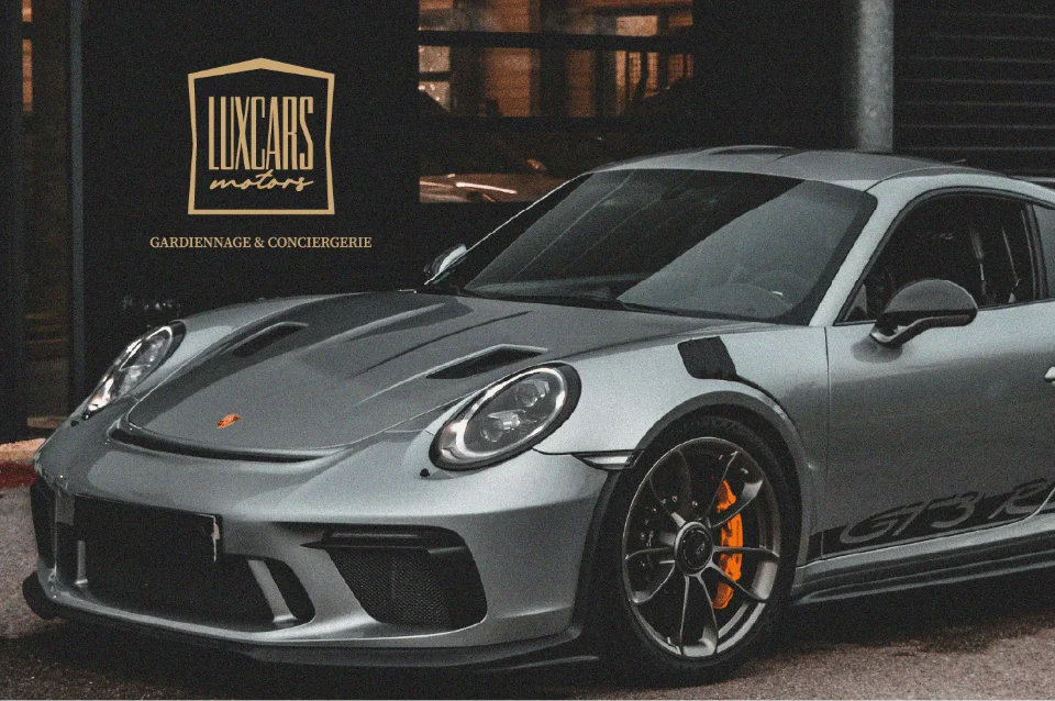 Porsche 911 GT3 RS grise aux étriers de frein orange, devant le logo Luxcars Motors.