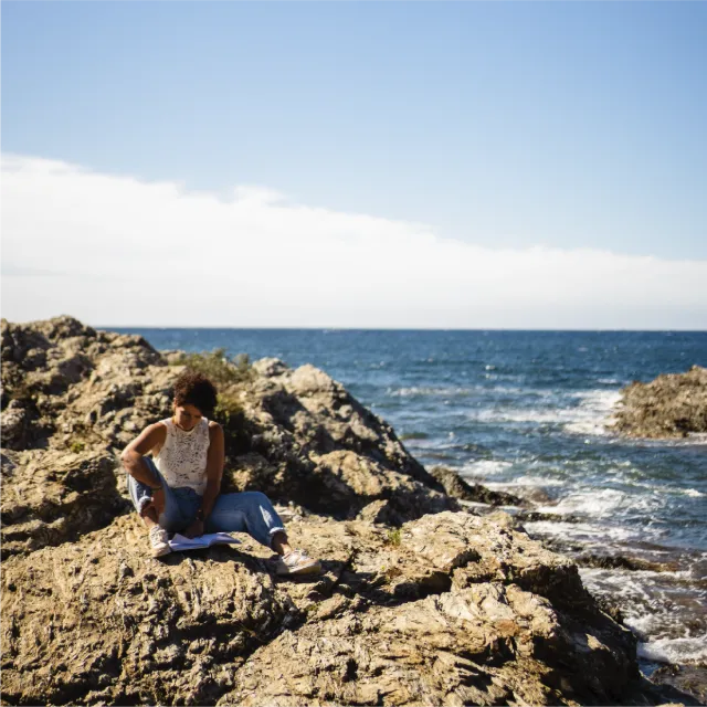 Graphiste La Tour du Pin - Femme aux cheveux bouclés assise sur des rochers au bord de la mer, regardant un livre ouvert sur ses genoux. Chloé Lance