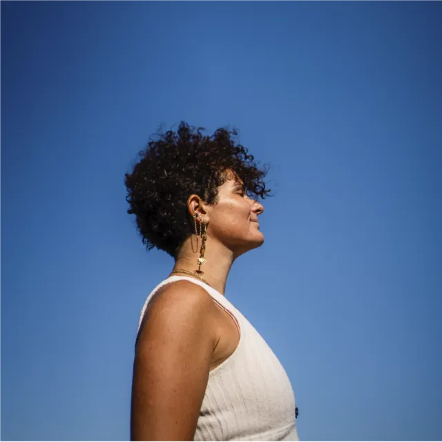 Graphiste La Tour du Pin - Profil d'une femme aux cheveux bouclés, portant une tenue blanche et des boucles d'oreilles originales, regardant vers le ciel bleu clair.